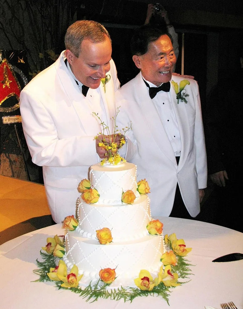 Two men in white tuxedos stand beside a three-tier wedding cake decorated with yellow roses, smiling together