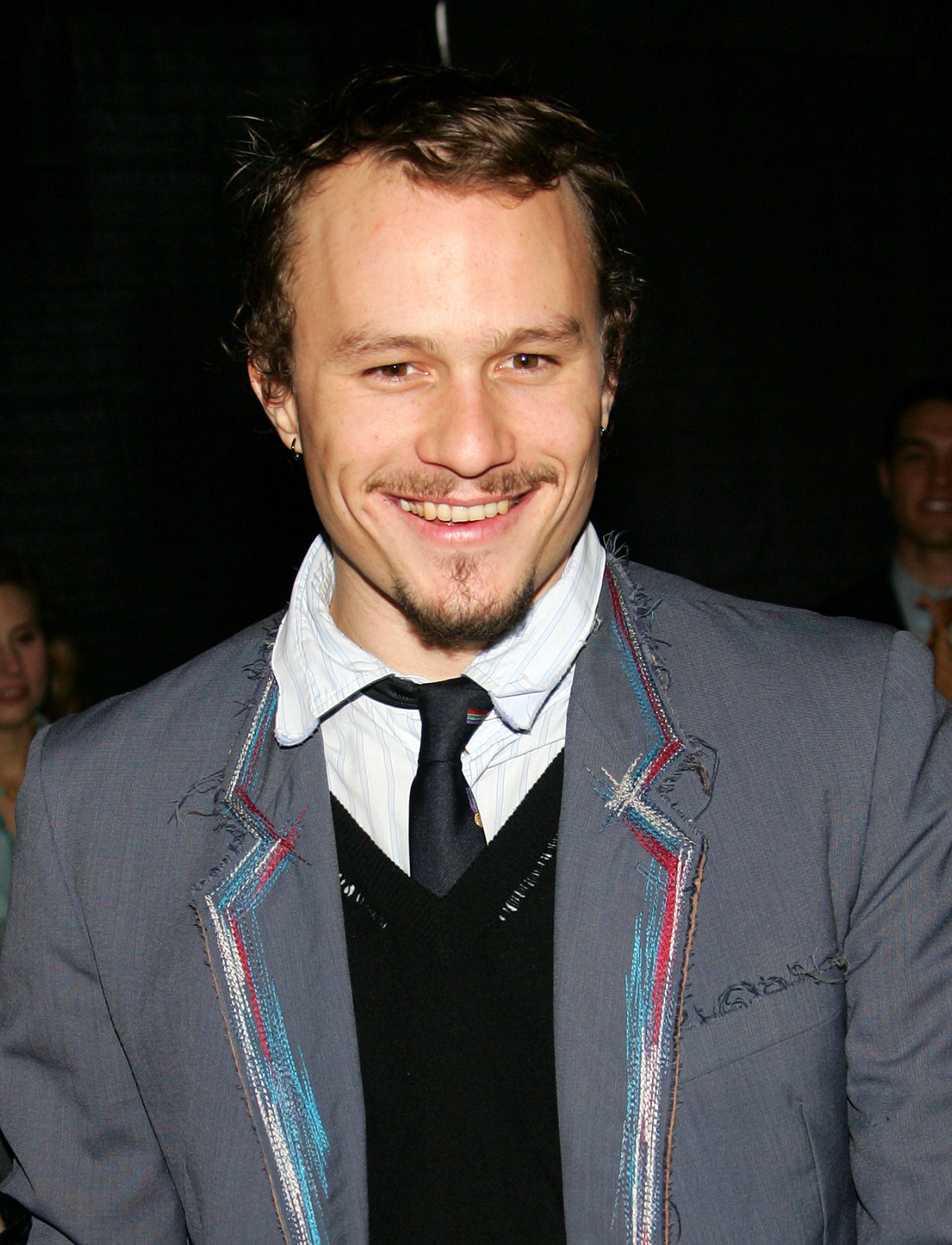 Heath Ledger wearing a suit with a black tie and smiling. The suit has embroidered edges