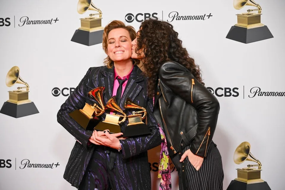Person in a patterned suit holding Grammy awards, smiling while another person in a leather jacket leans in to kiss their cheek