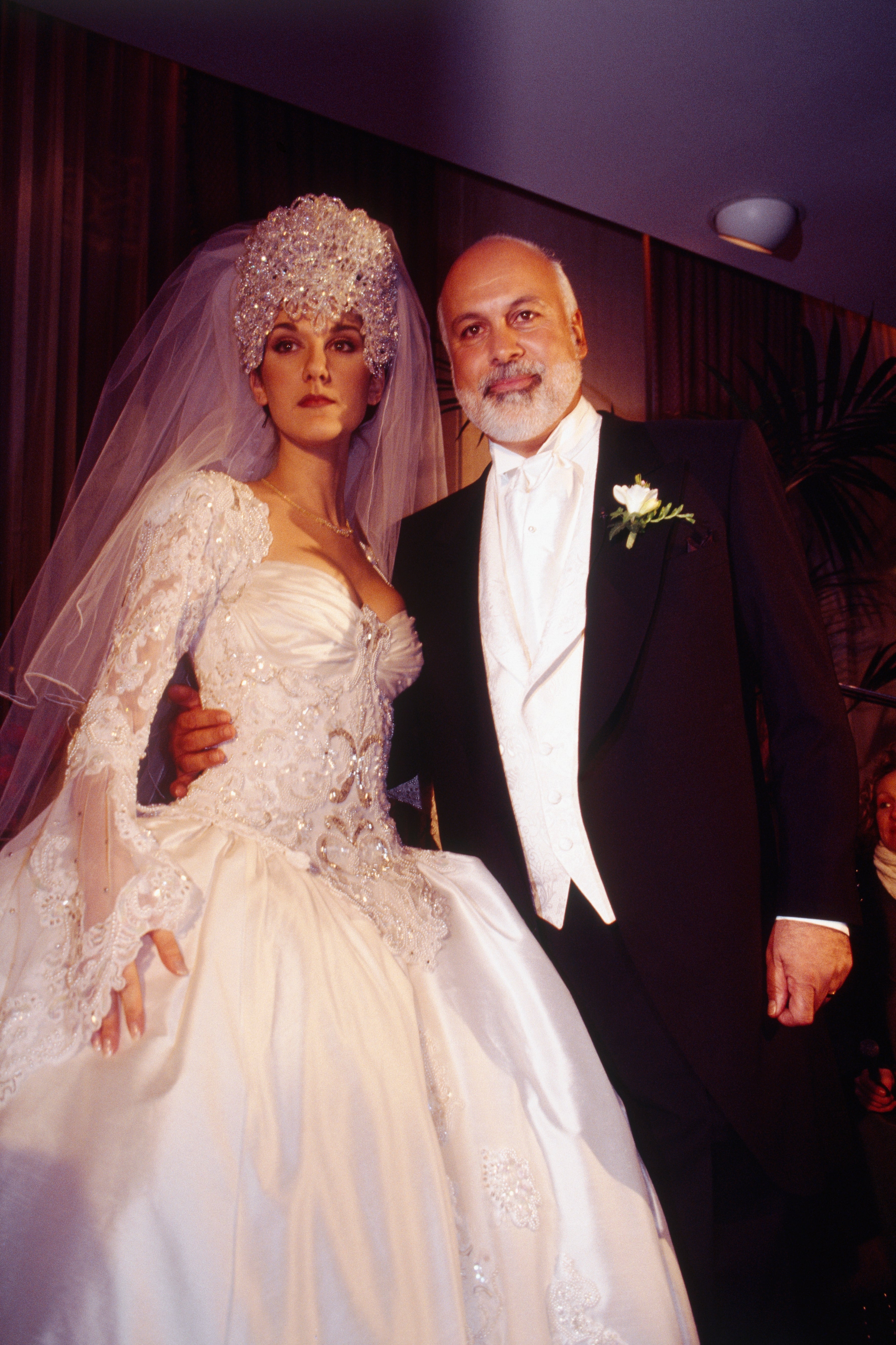 Bride in an elaborate wedding gown with lace and beading, posing with a bearded man in a formal suit, wearing a boutonnière