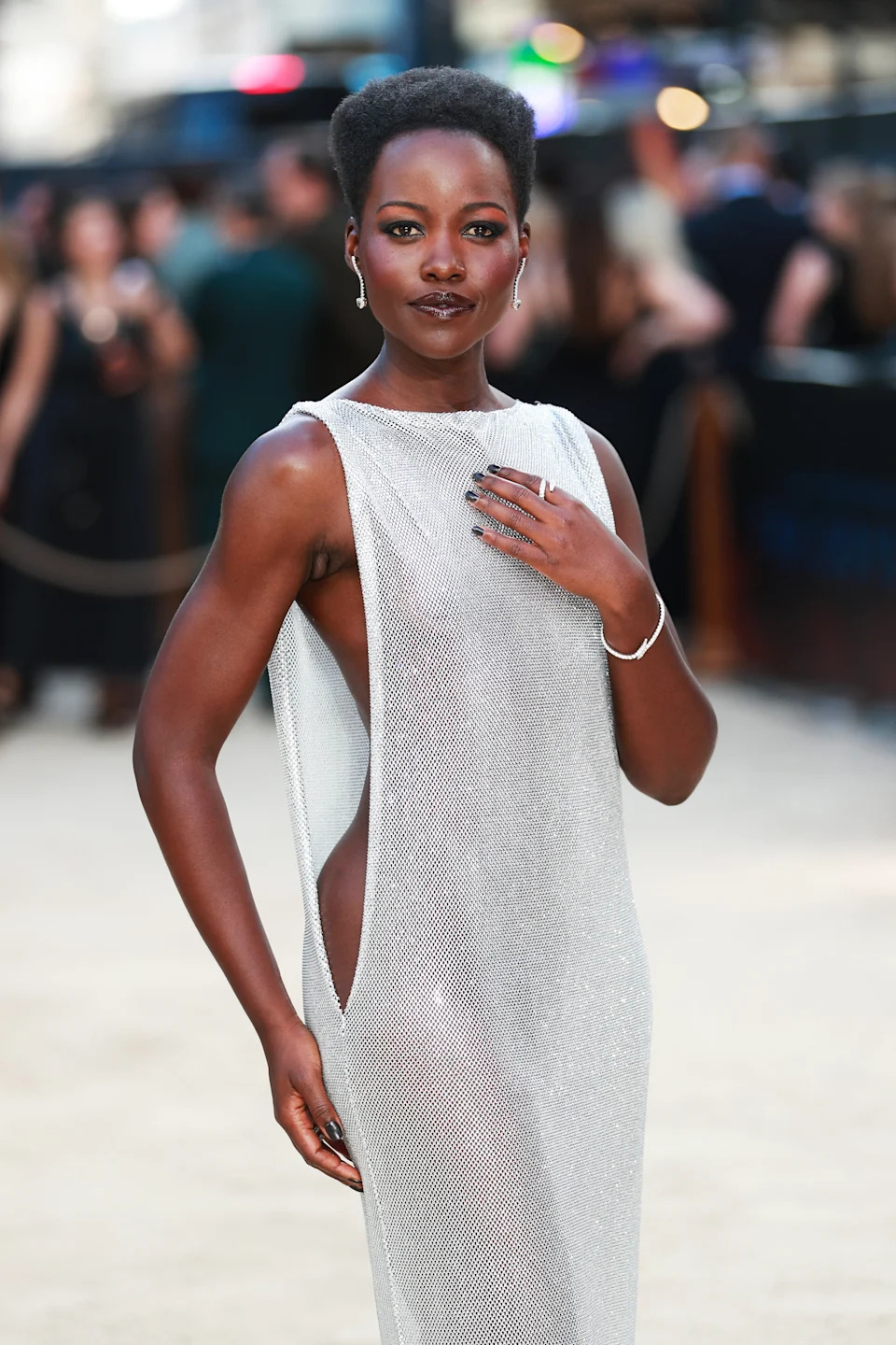 Person in a shimmering sleeveless gown on a red carpet poses confidently with elegant accessories