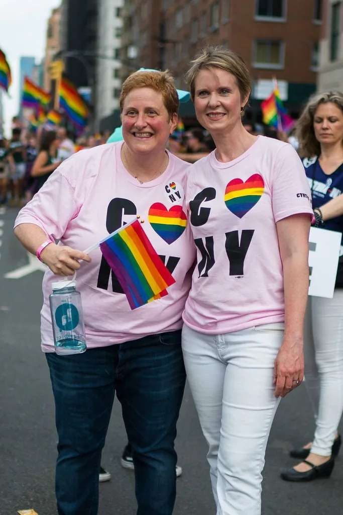 Two people smiling, wearing matching "G❤️NY" shirts, holding a rainbow flag, standing in a pride parade with rainbow flags in the background