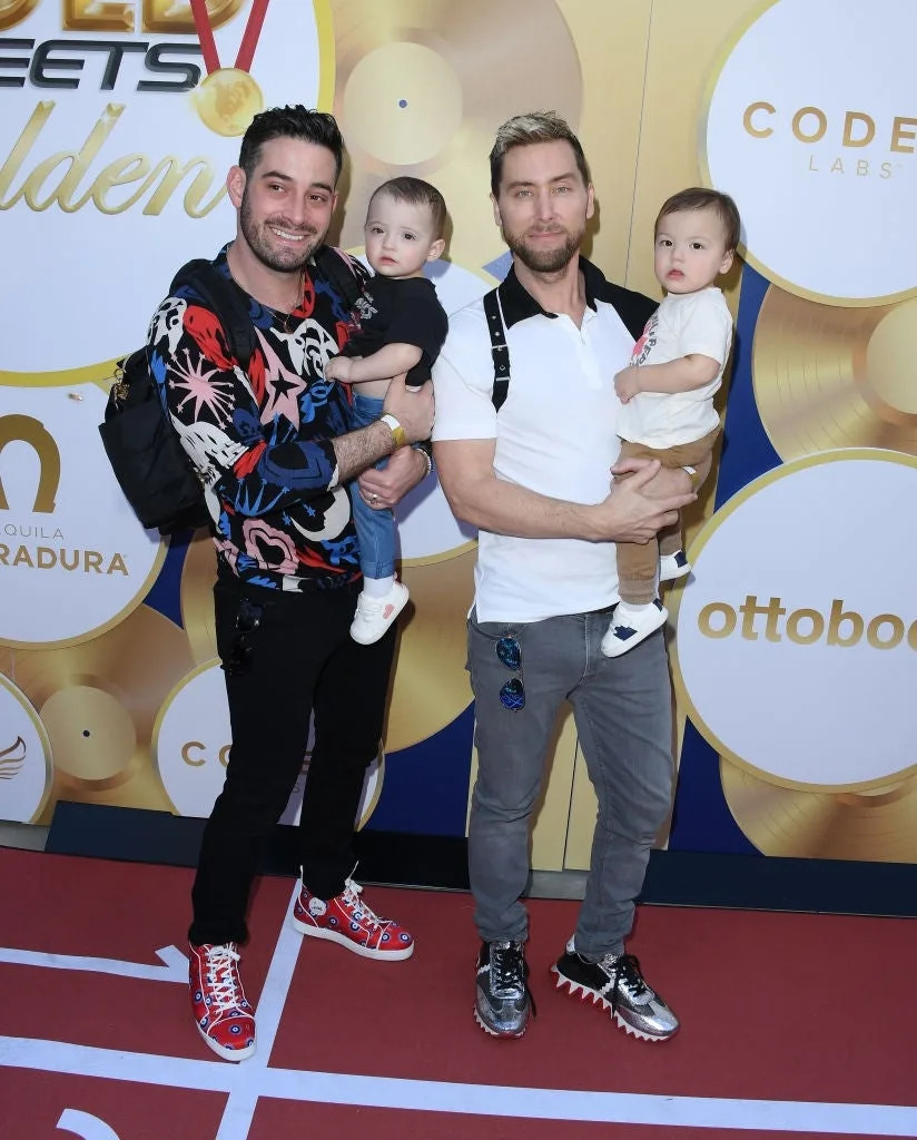 Two individuals on a red carpet hold babies. One wears a patterned sweater, the other a casual polo and jeans. The event backdrop has logos and gold records