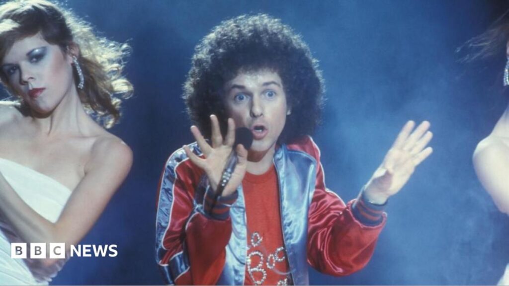 Leo Sayer speaks about growing up in Shoreham and West Sussex Beatles star Sir Paul McCartney smiles and waves from a car window while holding up a smartphone. Ringo Starr can be seen on the screen, wearing sunglasses. McCartney is dressed in a beige jumper with a light blue shirt collar underneath and several bracelets on his wrist.