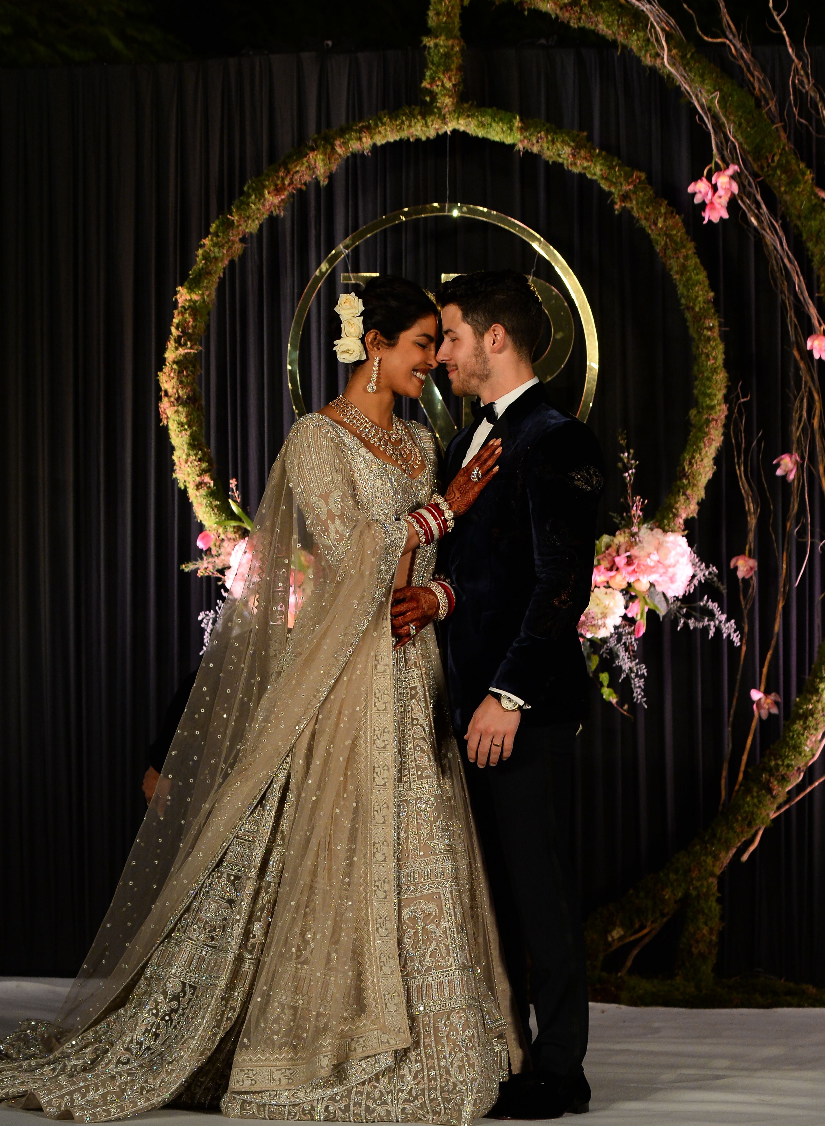 A couple stands closely, smiling, with the woman in an ornate, embroidered gown, and the man in a sleek suit, set against a floral backdrop
