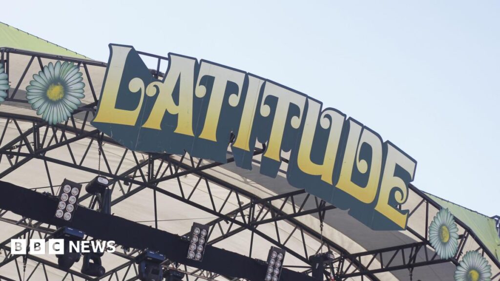 A close-up of the Latitude Festival sign on the top of a stage at the event. The letters are in block capitals, edged in black and coloured yellow fading to cream. Above it is sky.