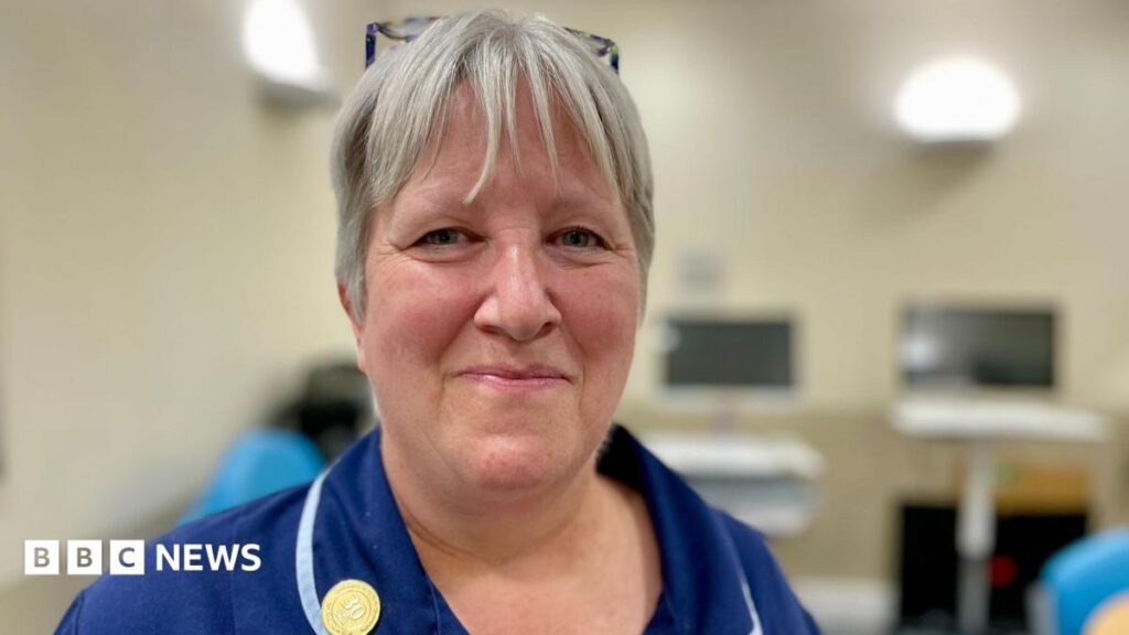 Kent hospital first to use AI to identify infections in patients A nurse in a blue uniform with ash blonde hair smiling at the camera standing in a clinical room in a hospital with computer screens in the background.