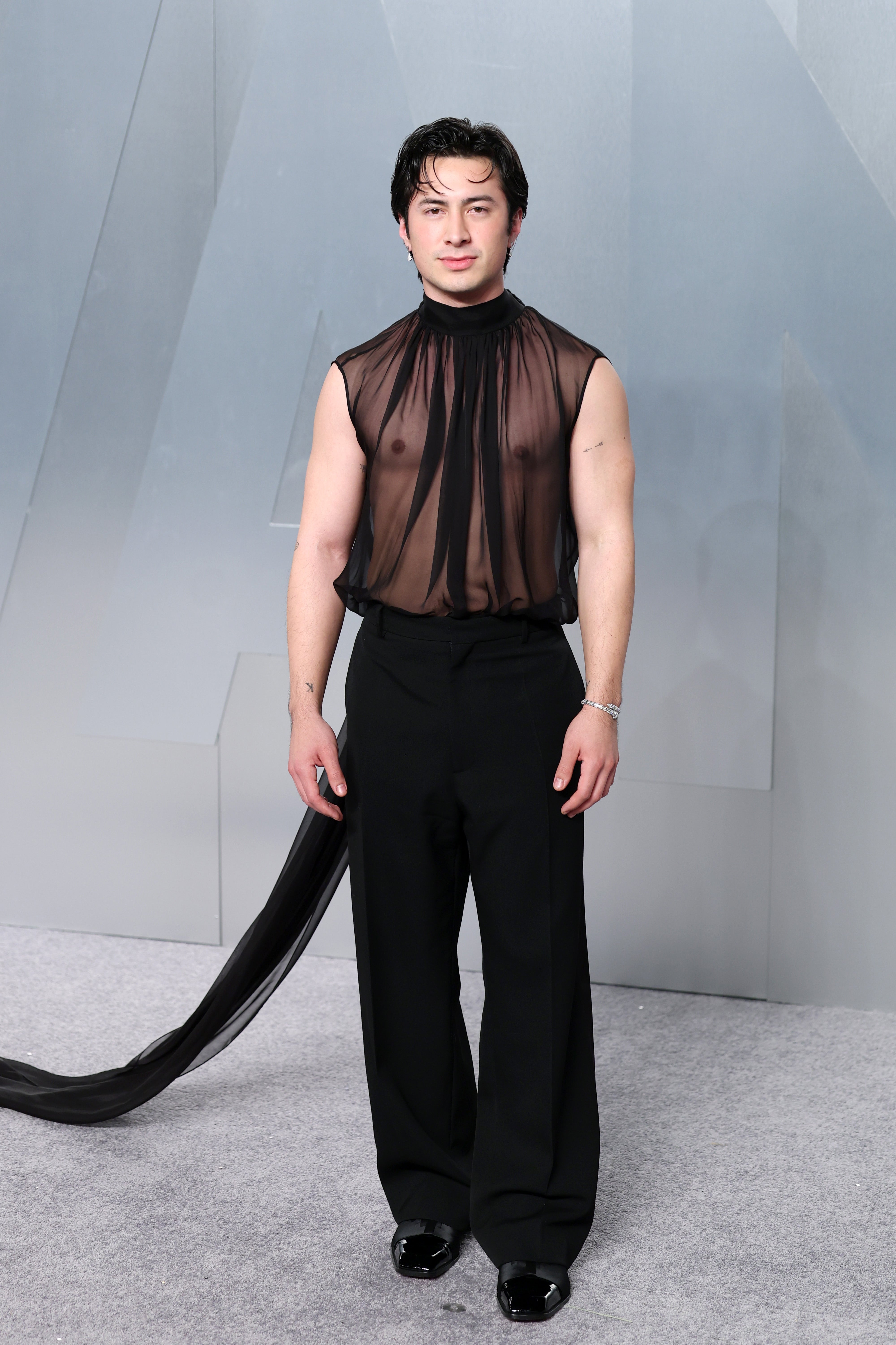 Person in sheer sleeveless top with flared black trousers and flowing scarf detail, standing on a light-colored carpet