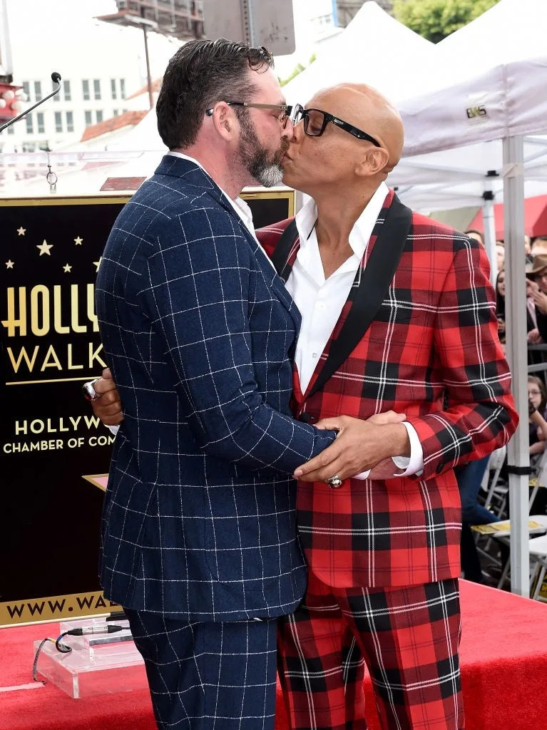 Two individuals share a kiss at a Hollywood Walk of Fame ceremony. One wears a blue check suit, the other a red plaid suit