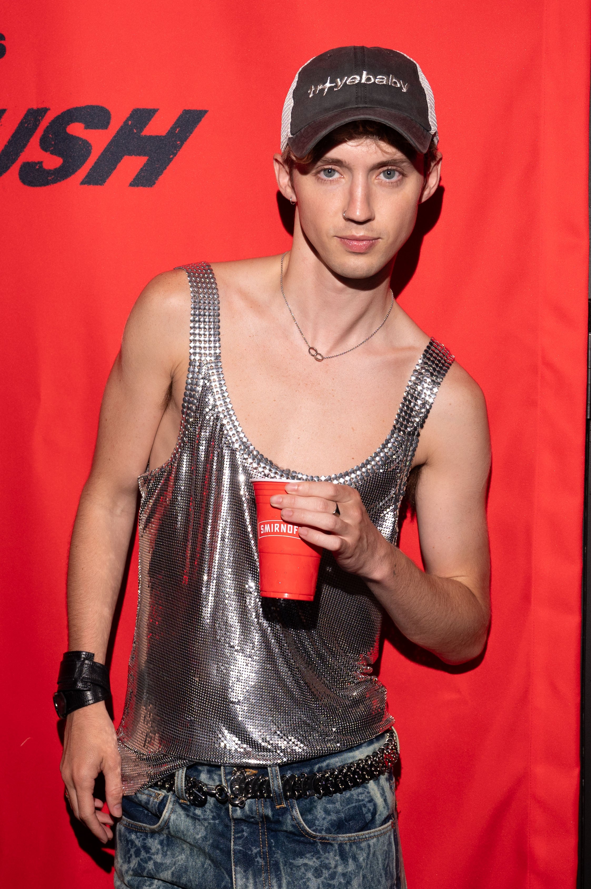 Person in a shiny tank top, jeans, and a cap, holding a red cup, stands against a red backdrop with text