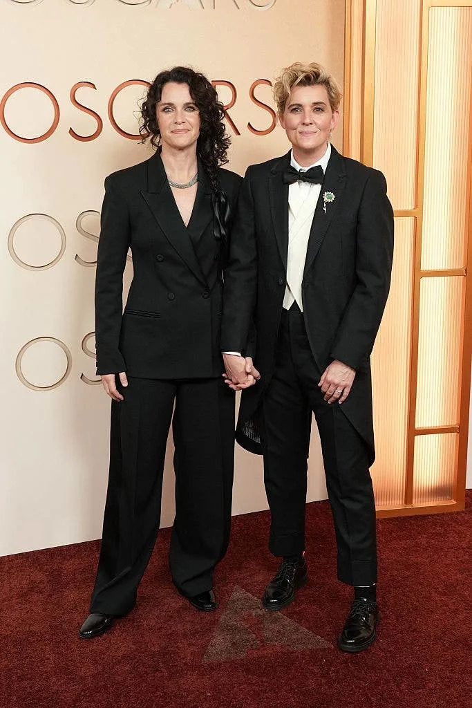 Two individuals on the red carpet. Left: black suit with a ribbon tie. Right: tuxedo with flower lapel. Both smiling and holding hands. Oscars backdrop