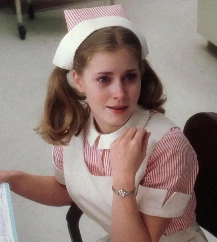 A young woman in a pink and white nurse uniform with pigtails sits on a chair, looking slightly concerned while holding a piece of paper.