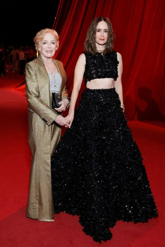 Two individuals on a red carpet; one in a shimmering suit, the other in a textured black crop top and skirt ensemble