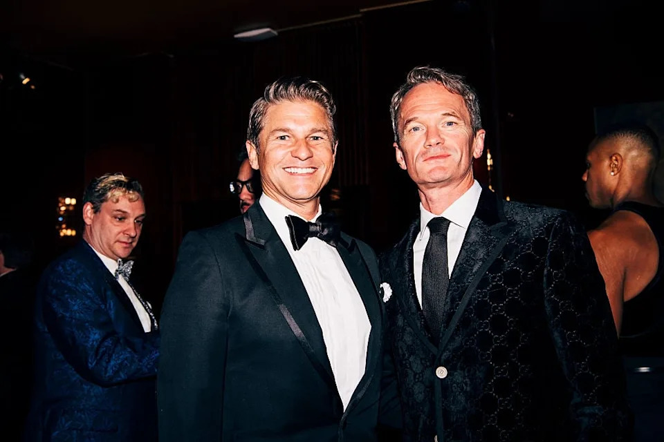 Two men in tuxedos smiling at an indoor event, attended by others in formal attire. One tuxedo has a distinct patterned texture