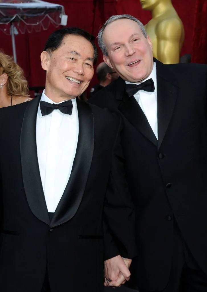 Two people in tuxedos smiling and holding hands on a red carpet, signifying a joyful public moment