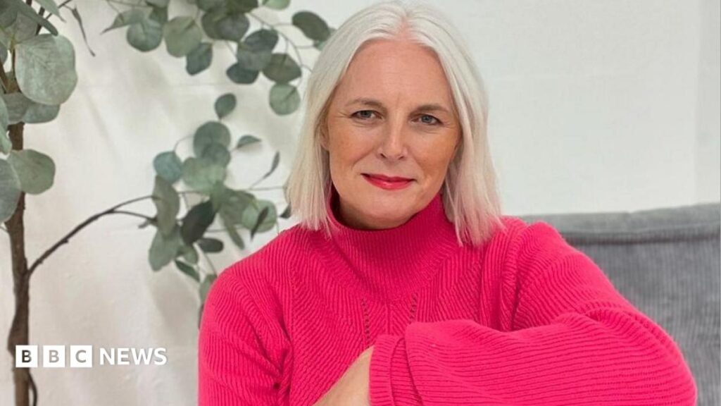 ‘My first big job modelling was alongside Lizzo’- Skipton model An woman with short white hair sits indoors on a grey cushioned seat, wearing a bright pink jumper and red trousers. A tall green houseplant in a woven basket stands behind them against a plain white wall.