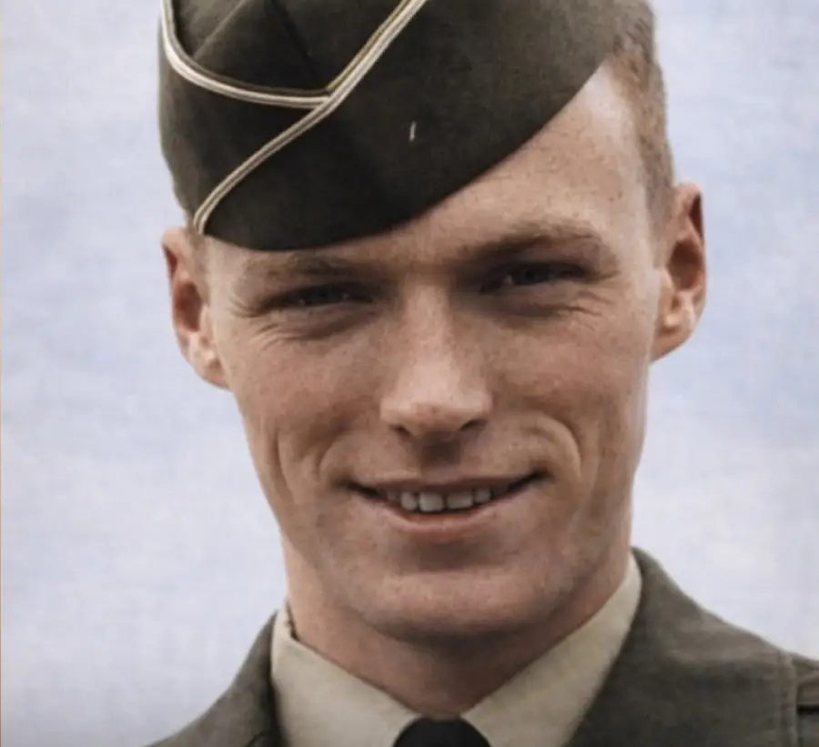 A young man in a military uniform and cap smiles at the camera against a pale blue background.