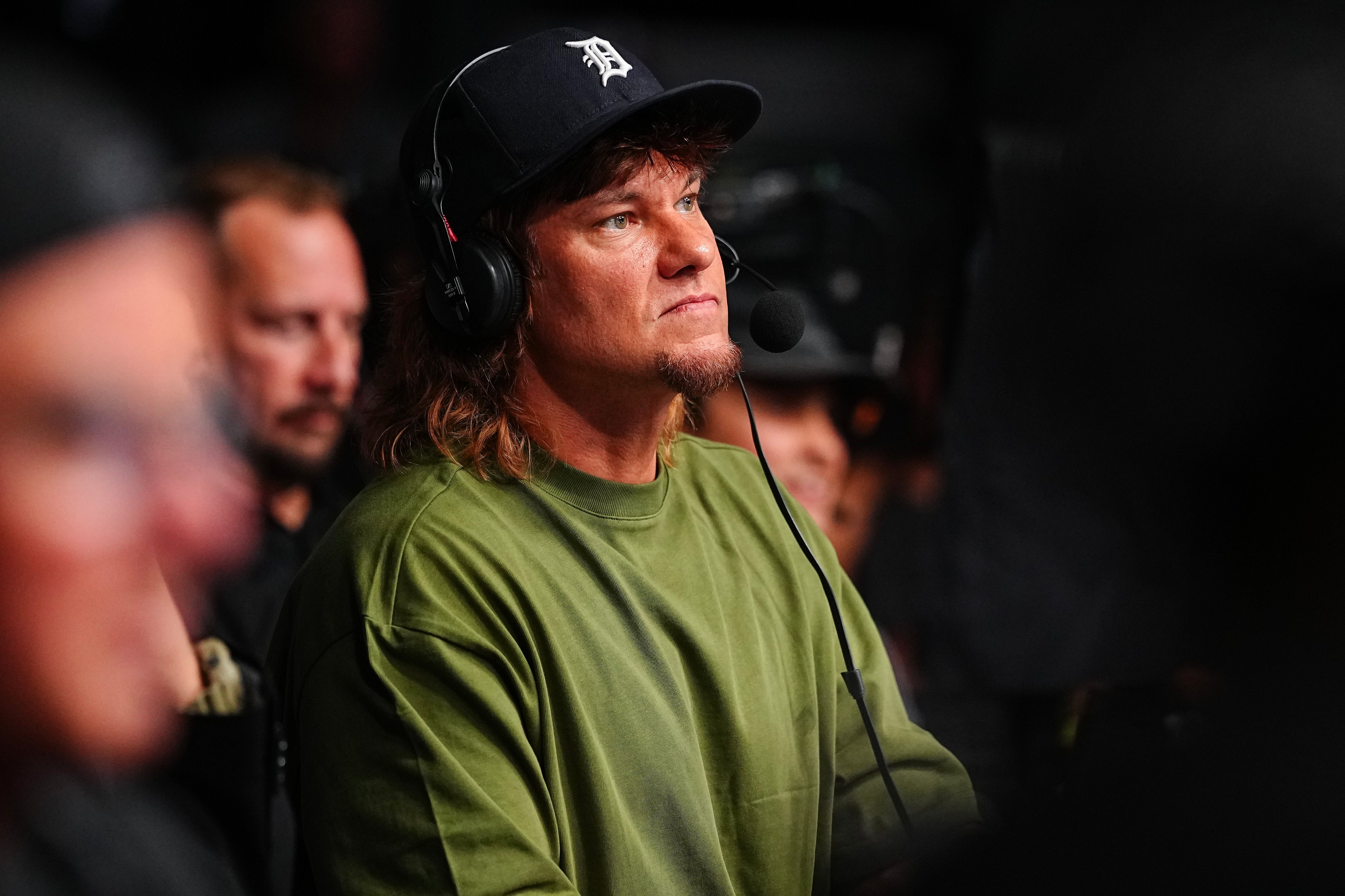 Theo Von wearing a cap and headset, seated and focused, with others in the background, at a UFC fight