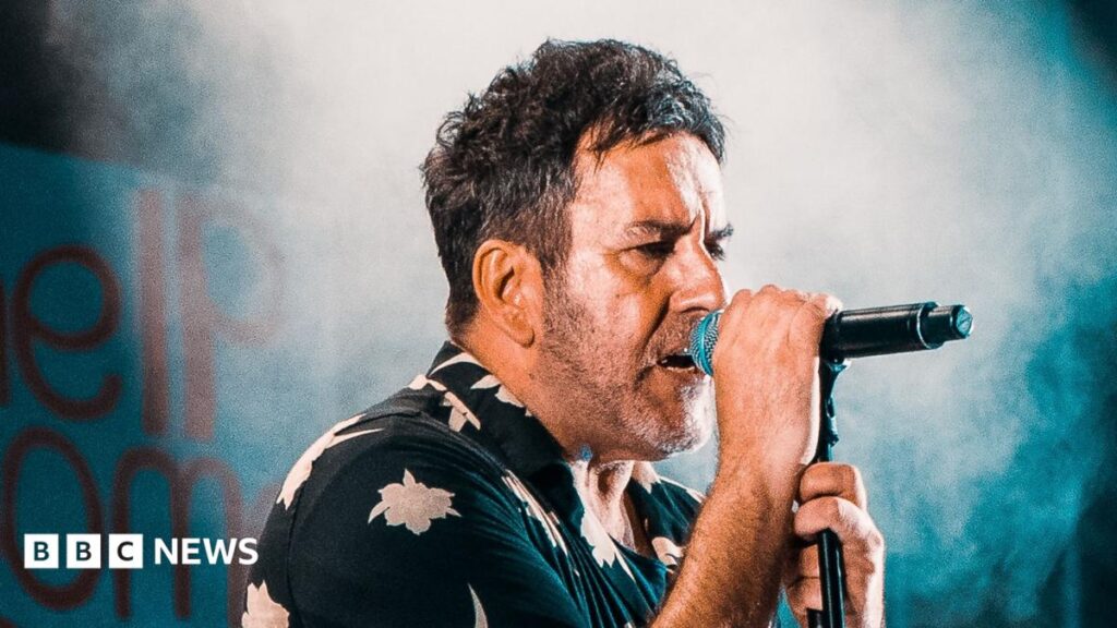The Specials’ last ever album ‘a fitting tribute to Terry Hall’ Terry Hall is wearing a dark shirt with white flowers on it. He is singing into a microphone during the 2019 concert at Coventry Cathedral