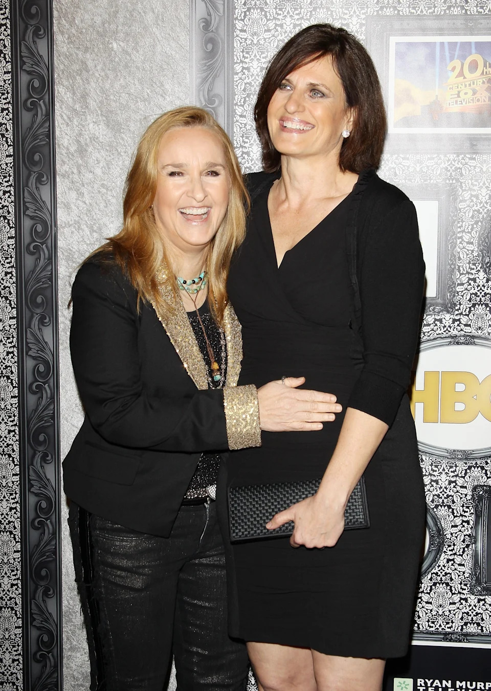 Two individuals smile for a photo at a formal event. One wears a casual black outfit with gold details, while the other is in a chic black dress