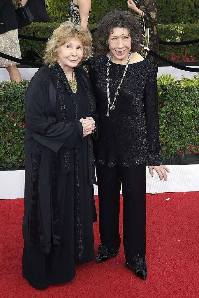 Two individuals on a red carpet; one in a long, flowing outfit, the other in a sleek, sparkling ensemble, both smiling at the event
