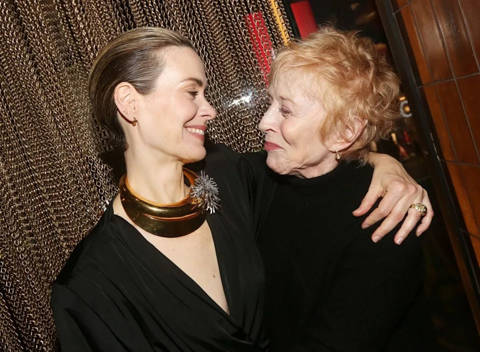 Two people warmly embrace, smiling at each other. The person on the left wears a sleek, black outfit with a bold necklace; the person on the right is in a black top
