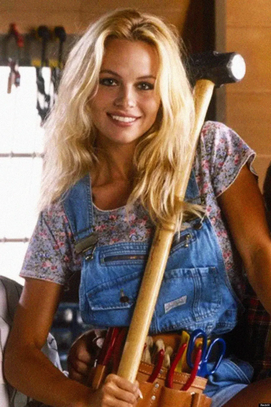 A woman with long blonde hair, wearing a floral shirt and blue overalls, smiles while holding a hammer. She has a tool belt with various tools, and is indoors with tools hanging on the wall in the background.
