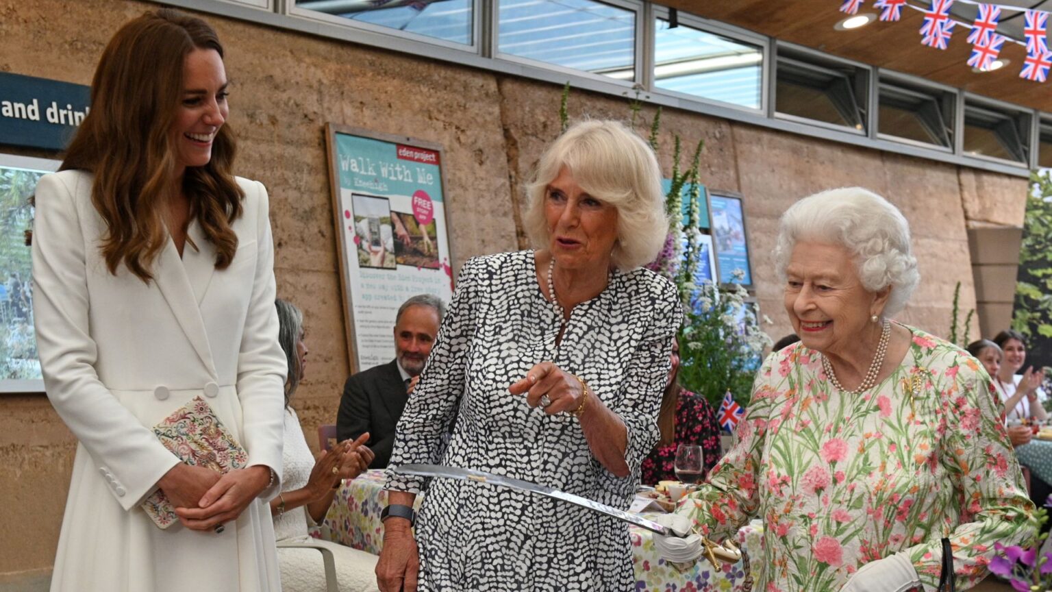 Queen Camilla Once Wore “Leathers” During a Motorcycle Phase Princess Kate wears a white coat dress and has long brown hair, she laughs as Queen Camilla, wearing a black and white patterned dress, points and Queen Elizabeth holds a large knife