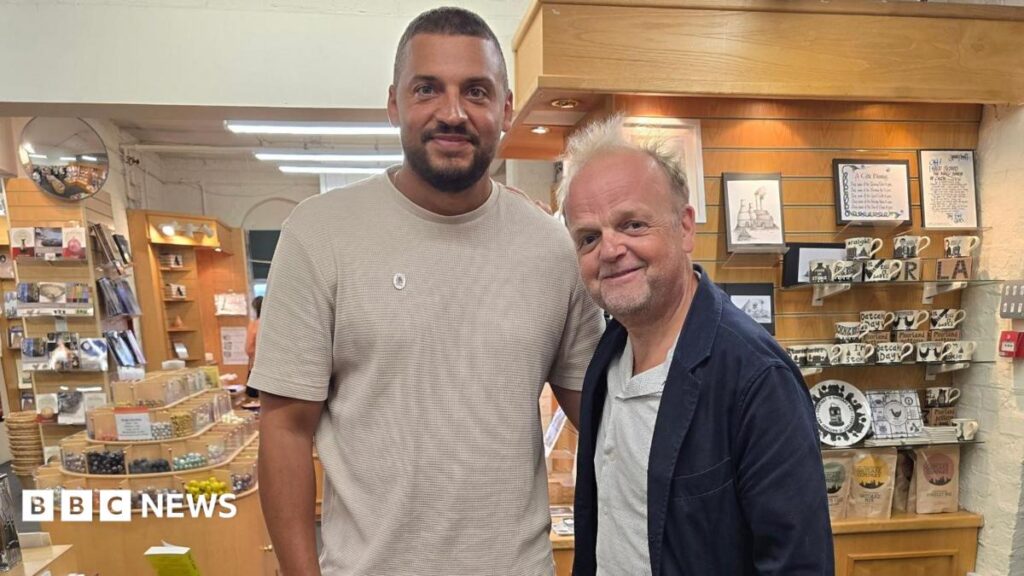 Ben Miller, a man with short dark hair, a beard and a beige shirt, stands next to actor Toby Jones, who is wearing a white shirt and dark blue jacket. They are both standing in a pottery gift shop.