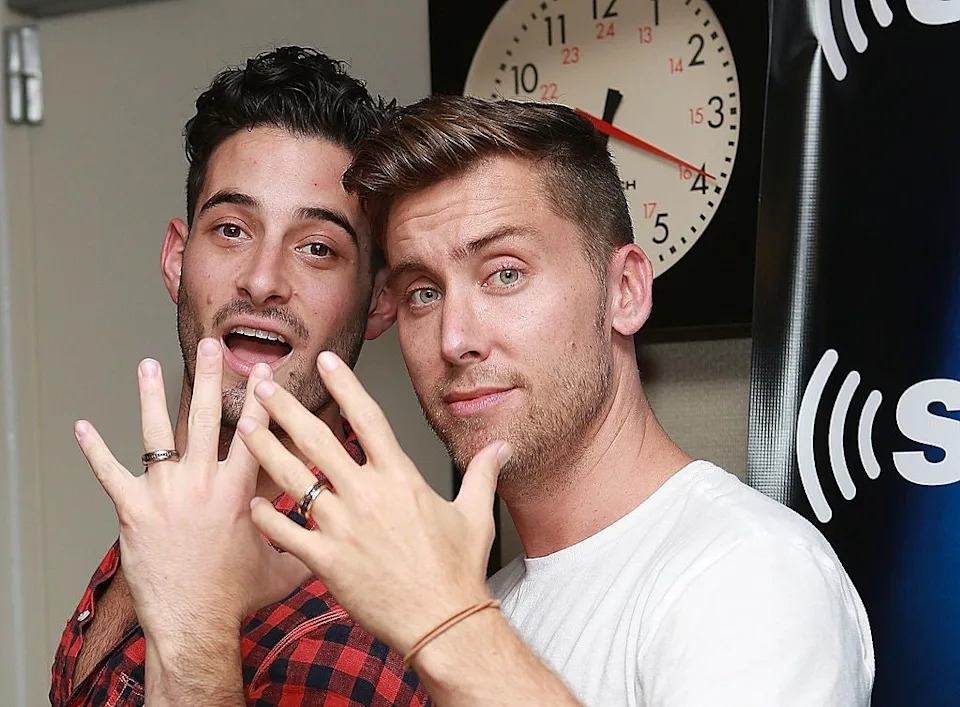 Two people smiling and showing rings on their ring fingers in a studio setting with a clock and a logo backdrop