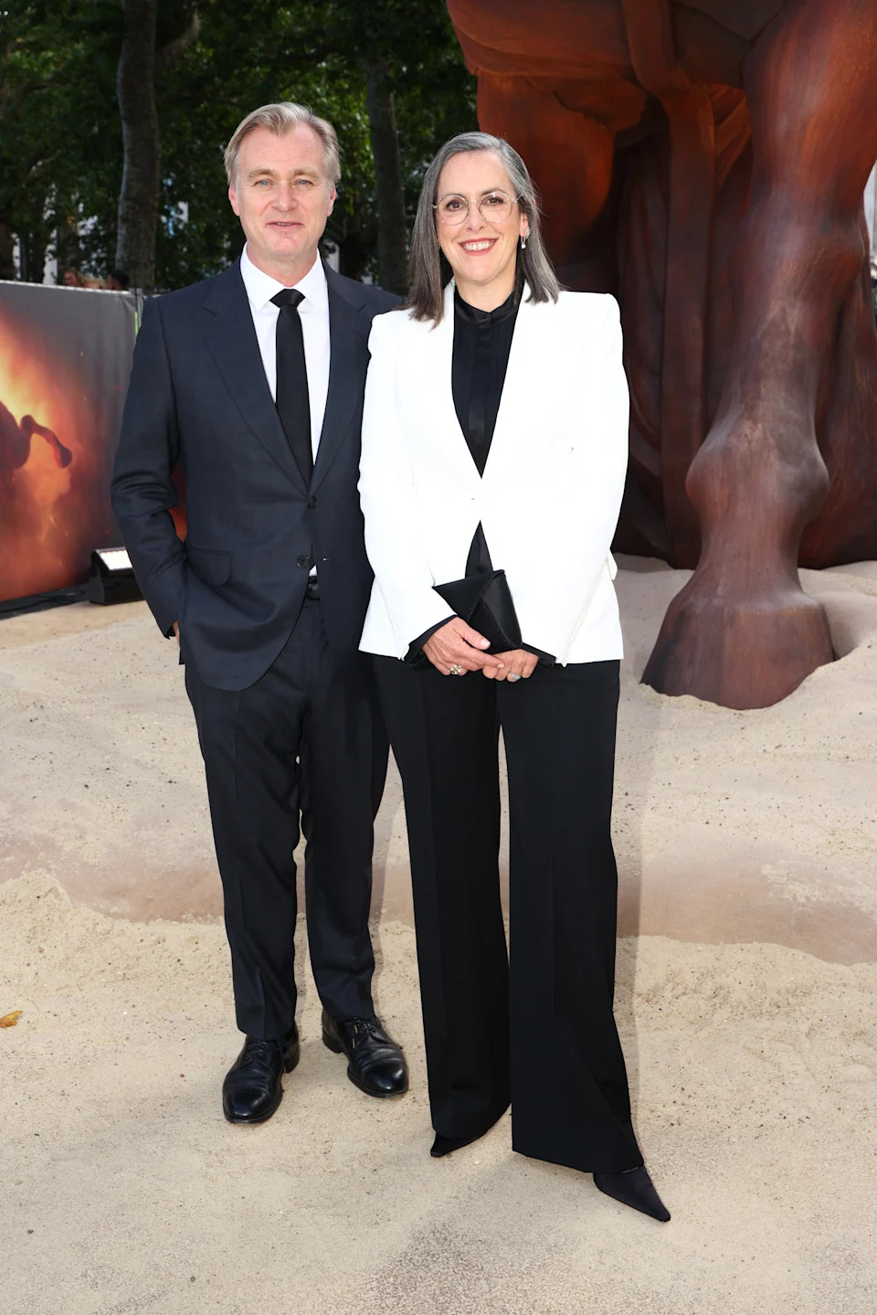 Two individuals pose on a sandy outdoor setting. The man wears a black suit and tie, while the woman wears a white blazer with black pants