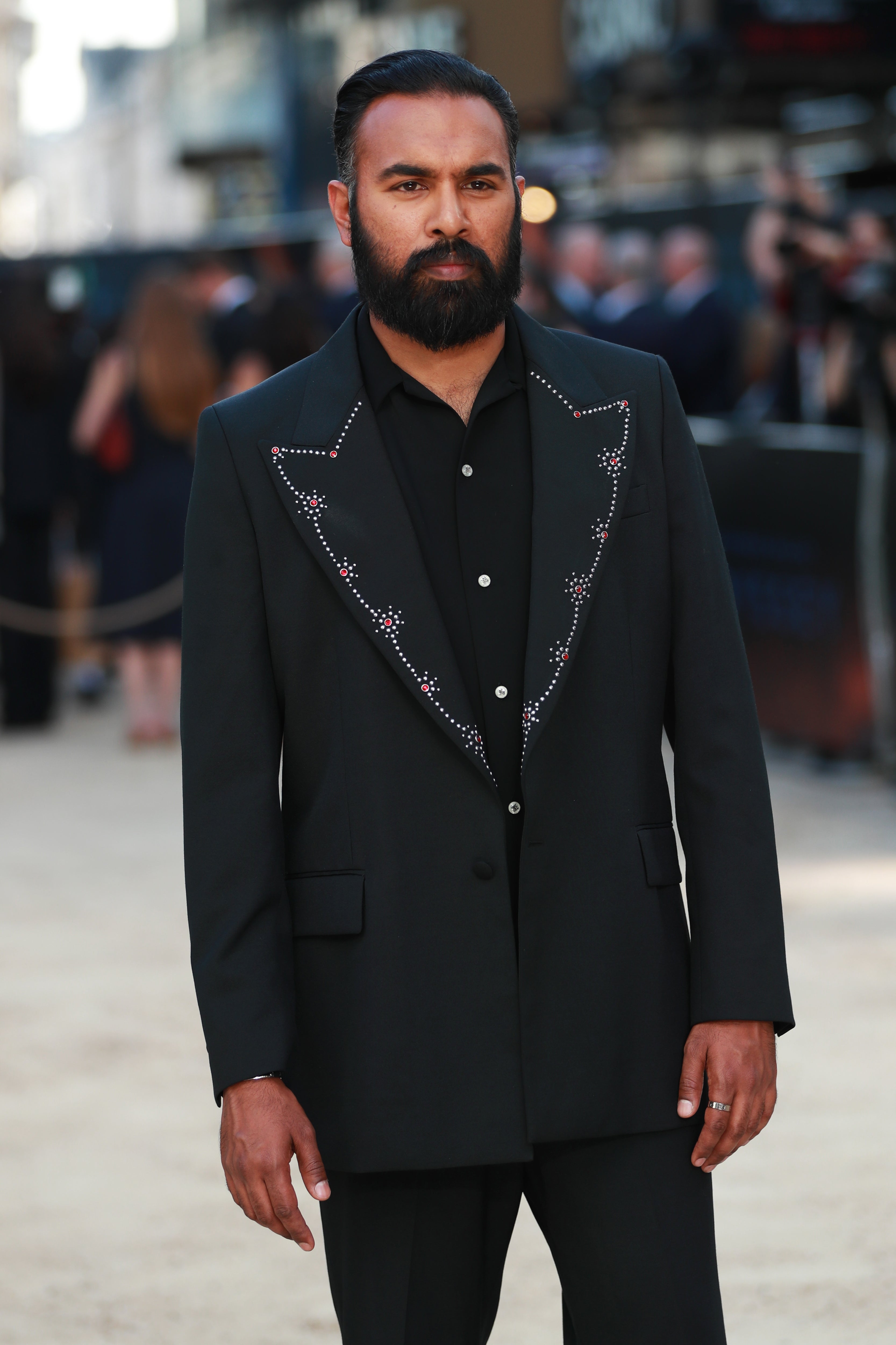 A person on a red carpet wears a stylish dark suit with beaded detailing on the lapels