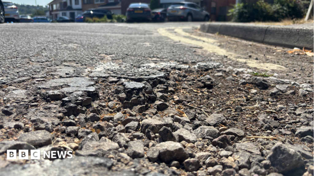 ‘Nightmare’ Cheltenham road has not been resurfaced for ’50 years’ A close up photograph of a pothole. There's lots of loose stones and patchy tarmac. There's two double yellows in the background next to the pavement. Out of focus at the top of the photograph is cars and houses from the street.