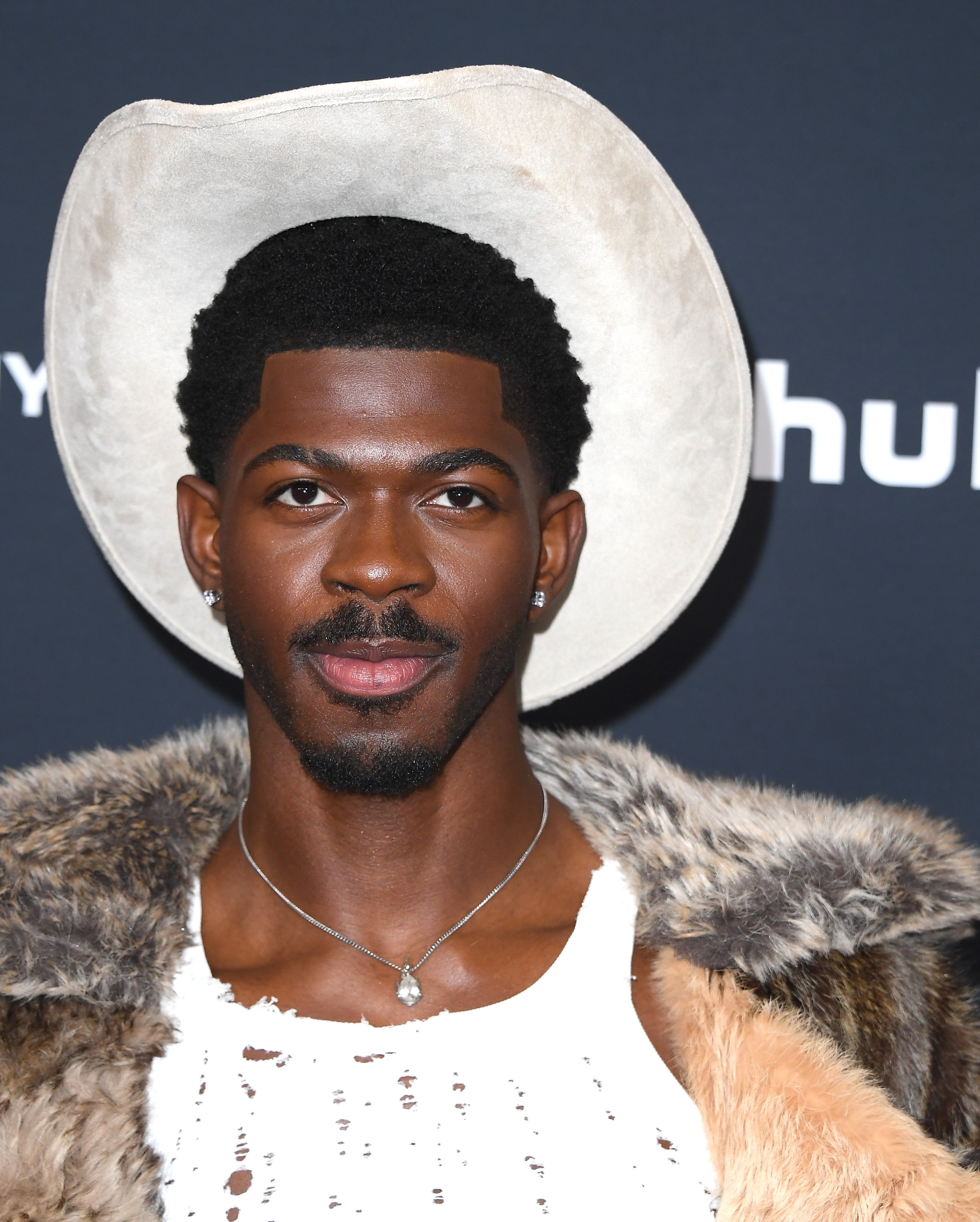 Person in a cowboy hat wearing a fur-trimmed coat and a torn, stylish top at a formal event