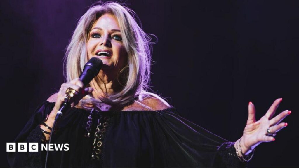 A glamorous woman with blonde hair, wearing all black and holding a microphone, with one hand raised up in the air.