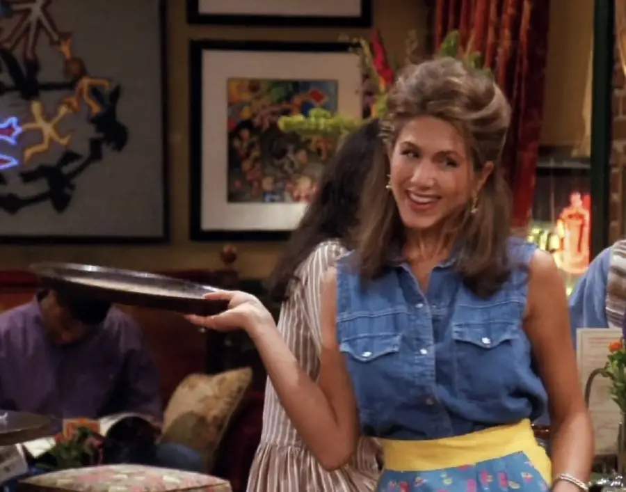 A woman with brown hair, wearing a sleeveless denim shirt and yellow apron, smiles while holding a serving tray in a colorful, cozy café setting.
