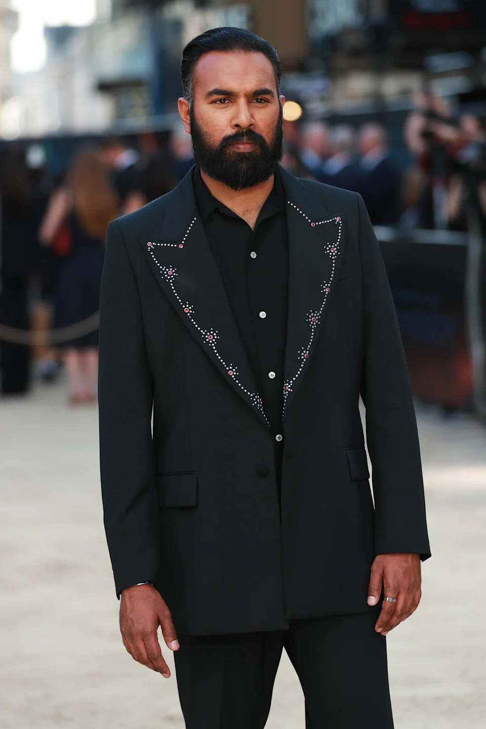 A person on a red carpet wears a stylish dark suit with beaded detailing on the lapels