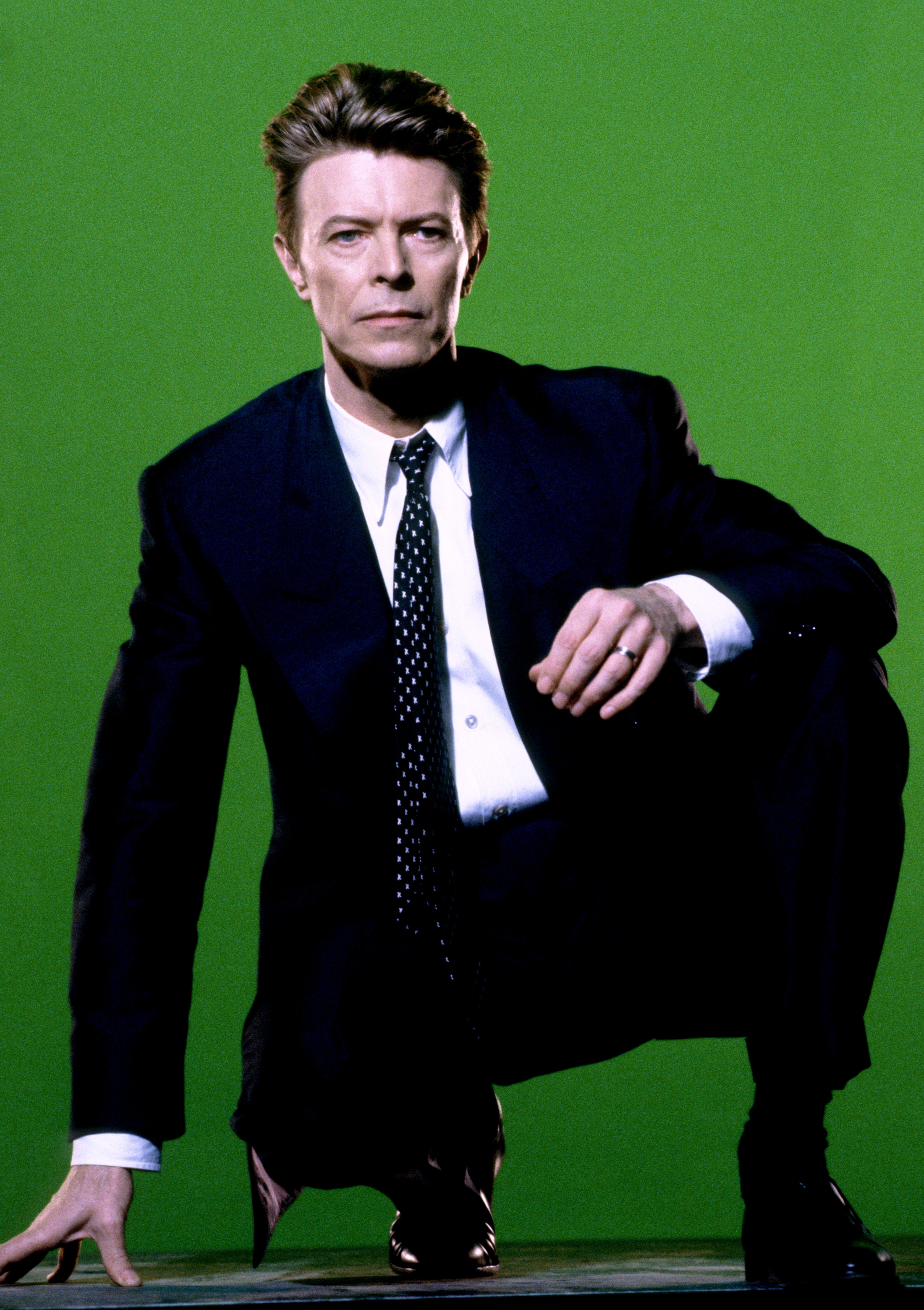 A person in a suit with a polka-dot tie crouches confidently against a plain background, exuding an elegant and poised demeanor