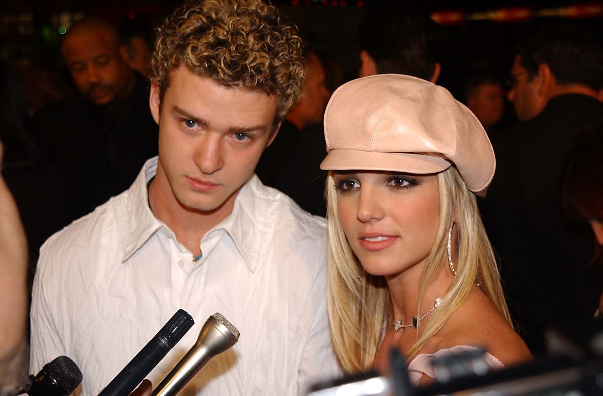 Justin Timberlake and Britney Spears stand side by side in front of microphones. Justin Timberlake wears a white, linen shirt, and Britney Spears wears a pink cap with large hoop earrings