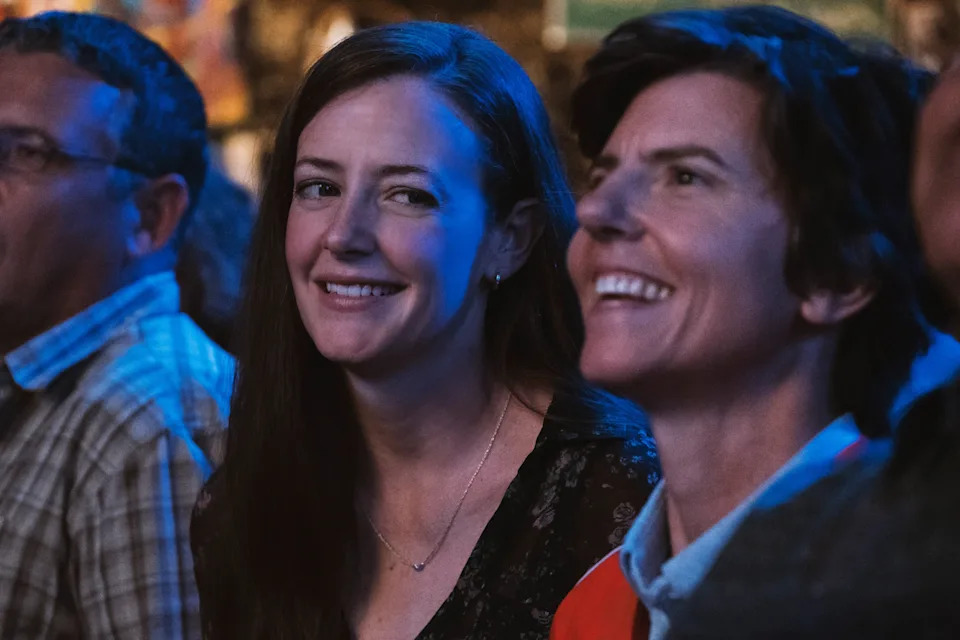 Two people smiling in a lively event setting, wearing casual outfits. The scene suggests a relaxed and joyful atmosphere