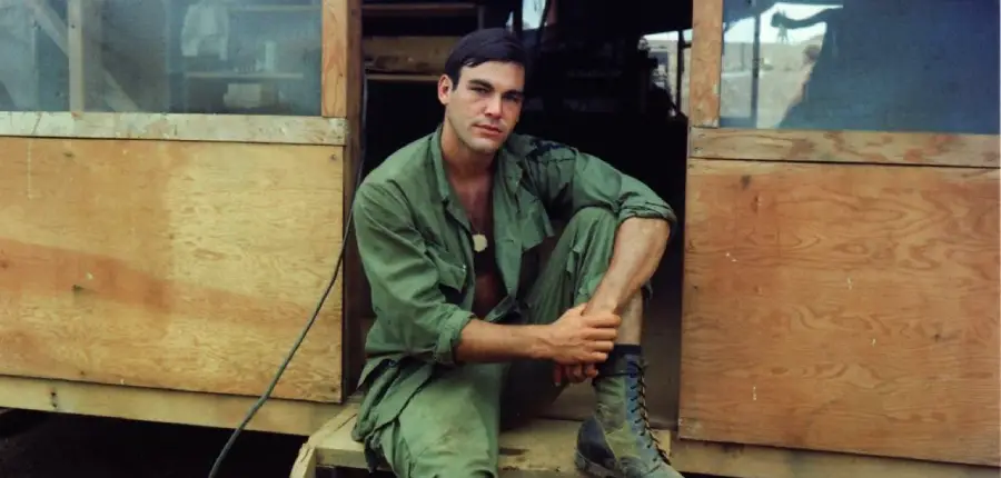 A young man in green military fatigues sits on wooden steps outside a wooden structure, looking at the camera with a relaxed expression.