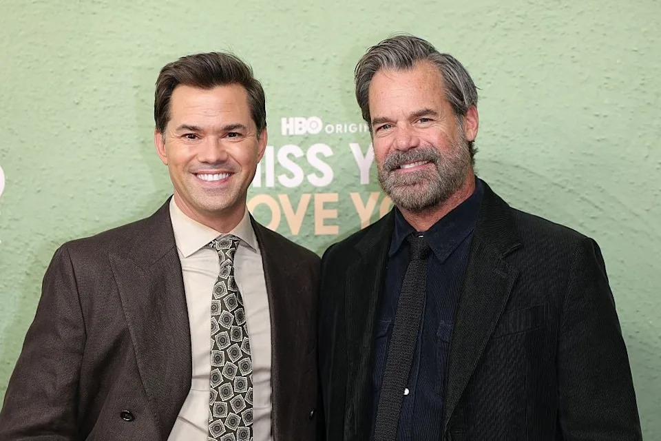 Two people smiling at an event. One person is in a brown suit with a patterned tie, the other in a dark jacket and shirt. Background has text