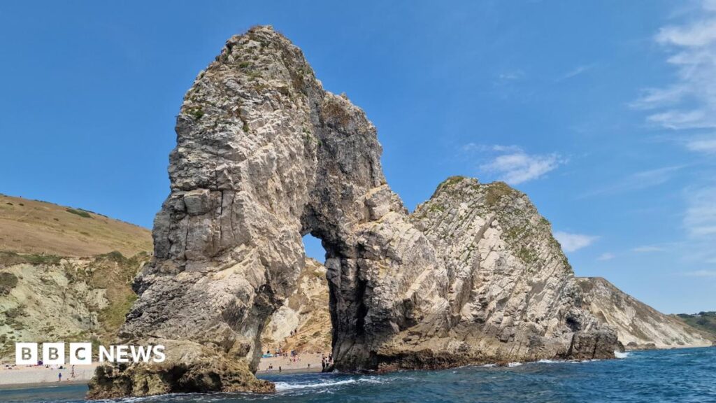 Big Picture: This week’s images from the south of England A large rock cliff arch sticks out into the sea. People can be seen on the beach through the arch. There are cliffs behind topped with browning grass. The sky is blue.
