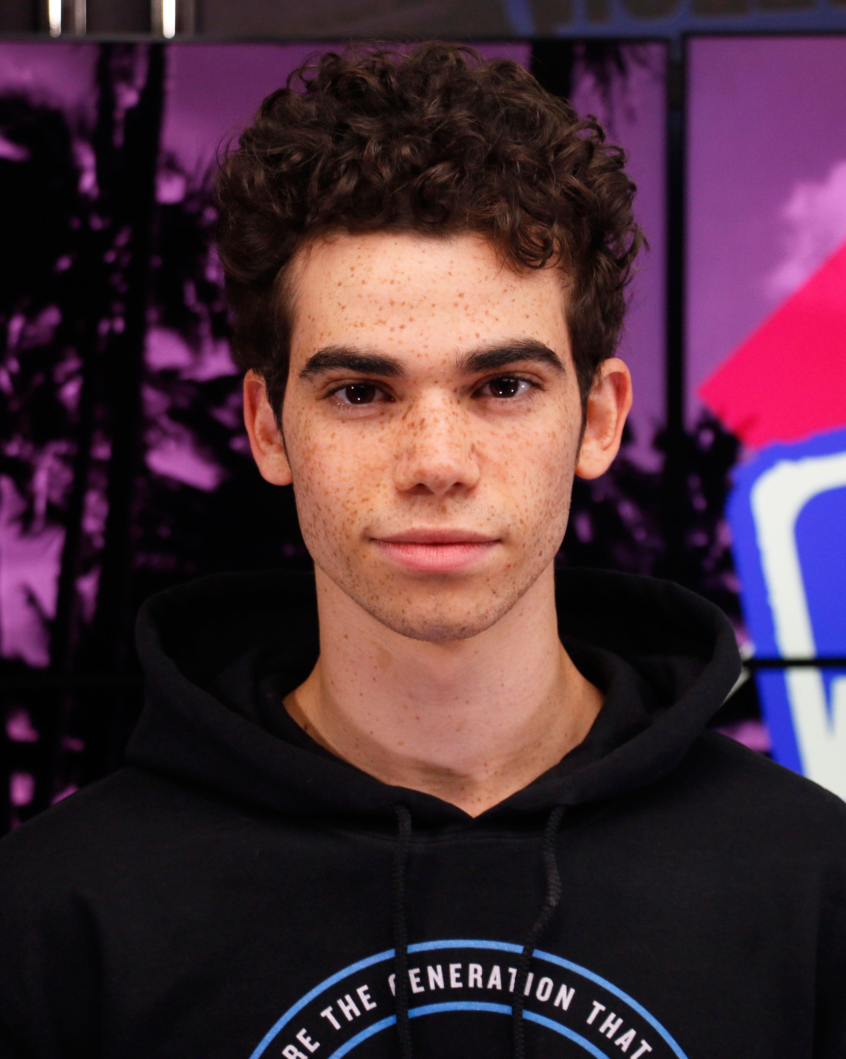 Cameron Boyce  with curly hair, wearing a hoodie, posing in front of a colorful backdrop
