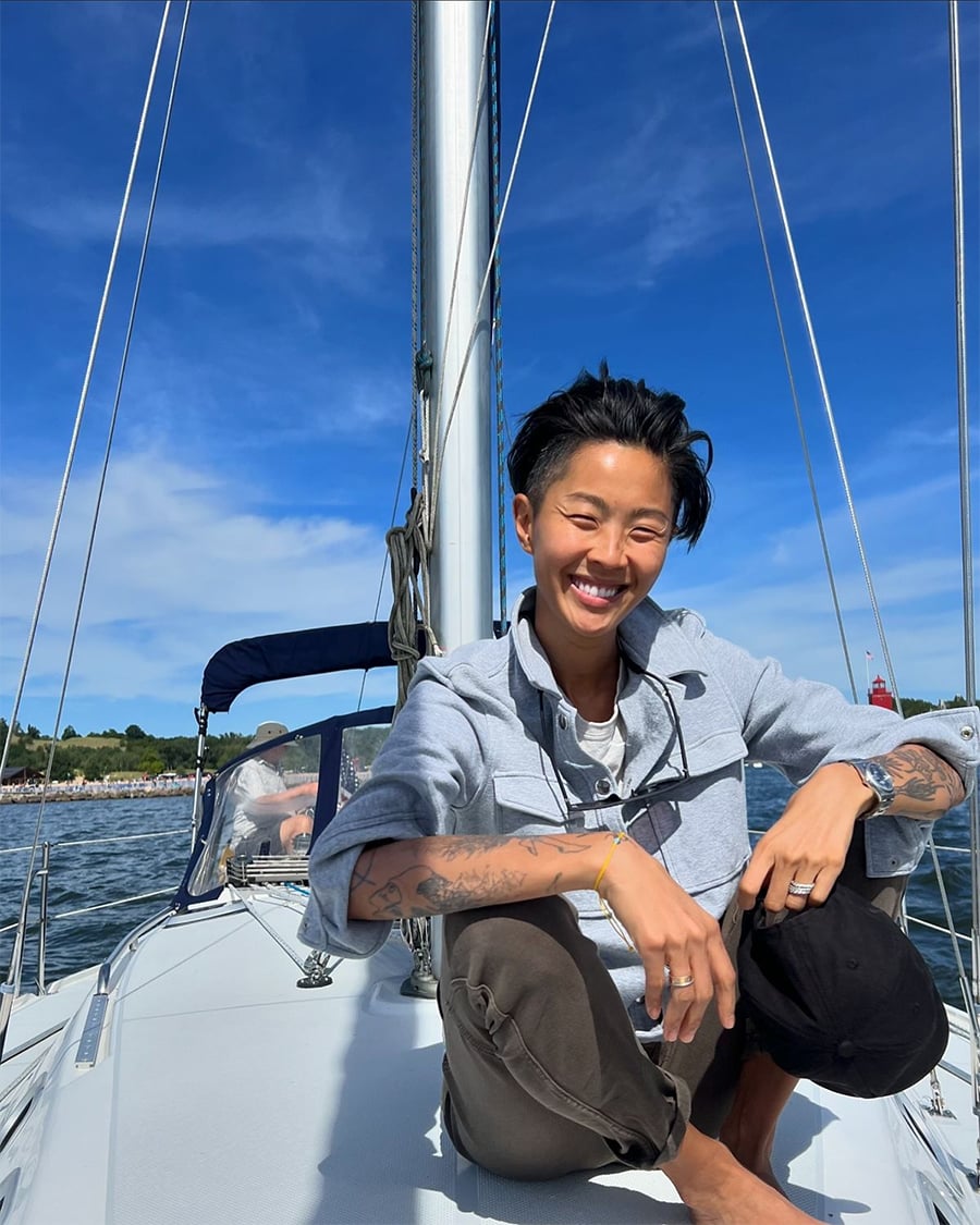A person with short hair and tattoos smiles while crouching on the deck of a sailboat on a sunny day, with blue sky, water, and green shoreline in the background.
