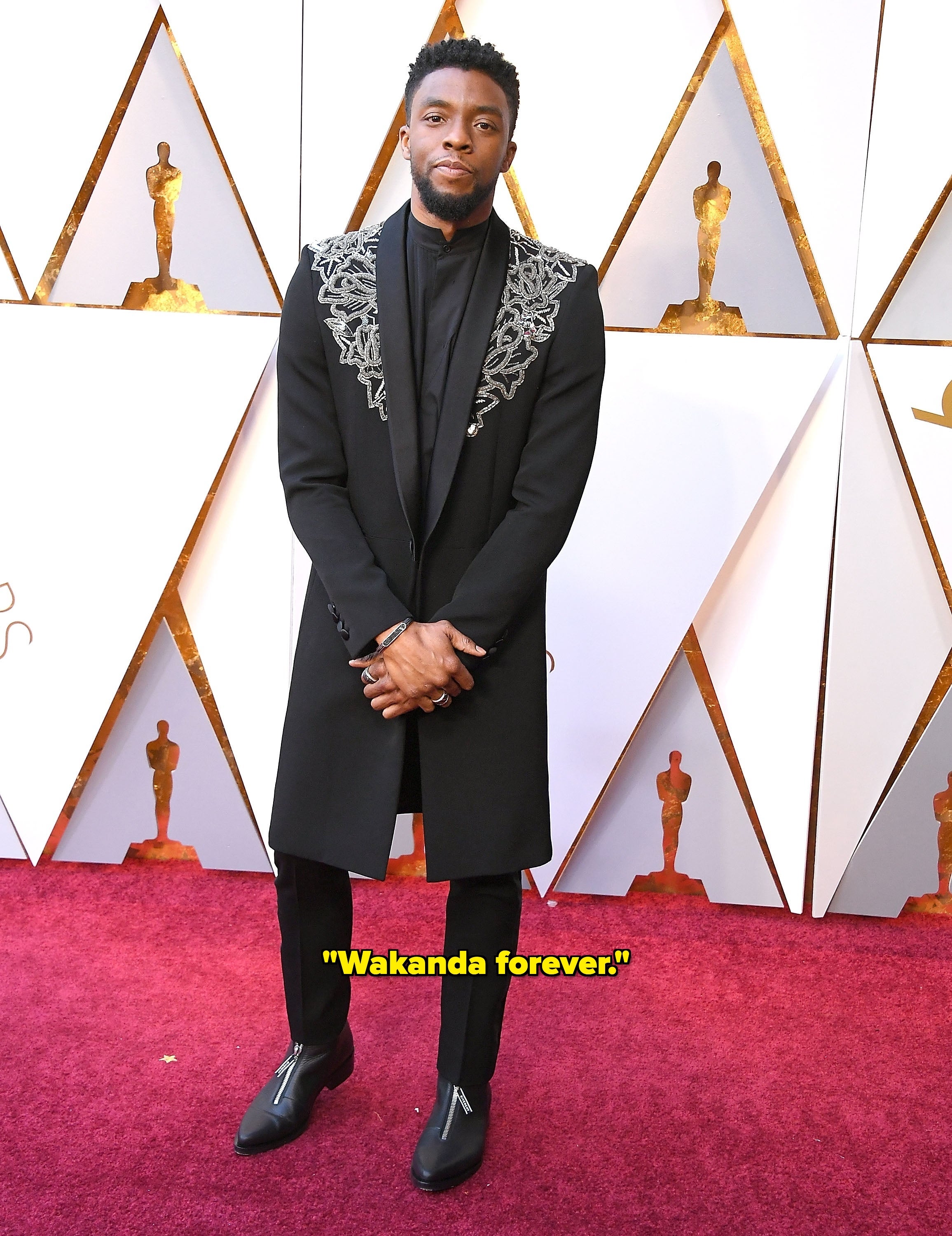 Chadwick Boseman in a stylish black suit with intricate embroidery stands on a red carpet