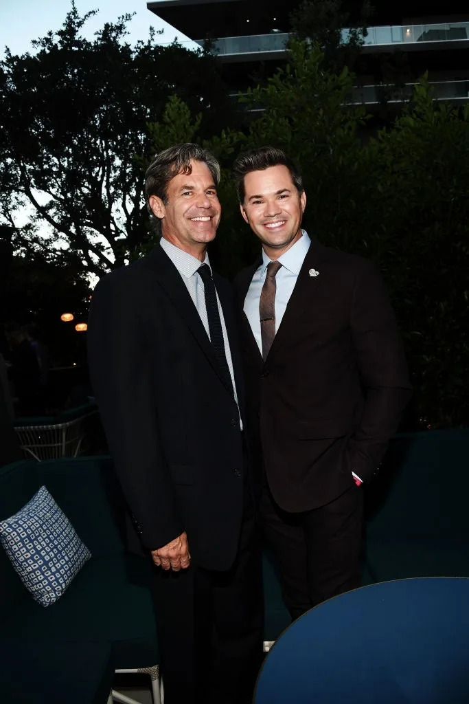 Two people in formal suits smiling outdoors at an event. The setting suggests a casual, upscale gathering with trees and soft lighting in the background