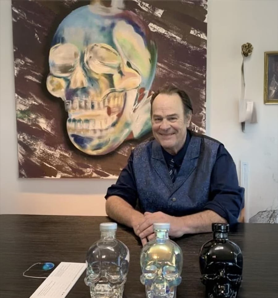A man in a blue vest sits at a table with three skull-shaped bottles in front of him. Behind him hangs a colorful painting of a skull on the wall.