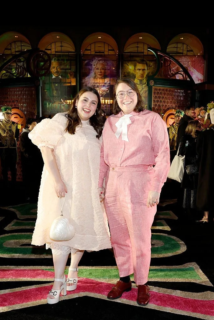 Two people on a red carpet, one in a lace dress with puff sleeves, the other in a suit with a white bow. They are smiling and holding hands