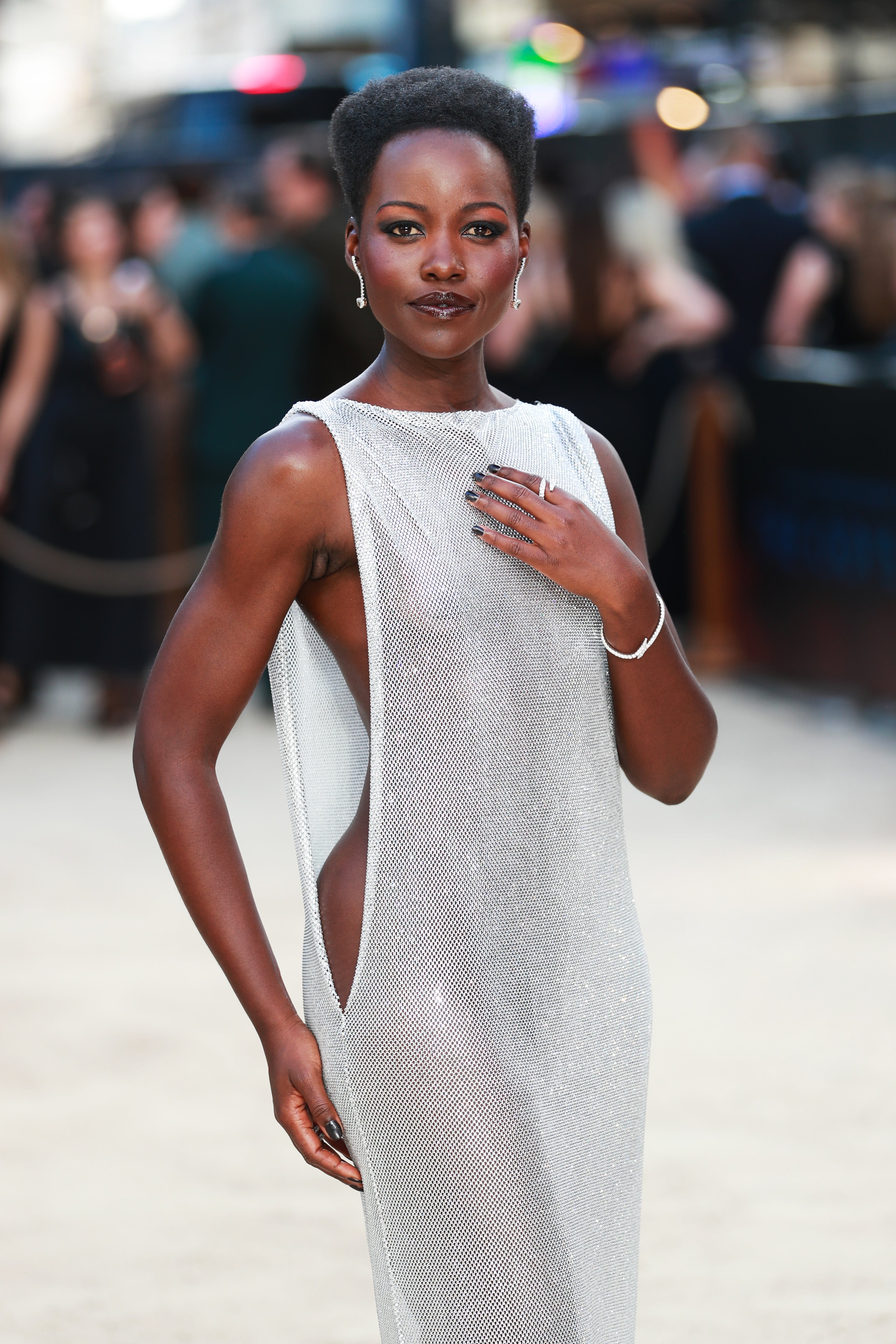 Person in a shimmering sleeveless gown on a red carpet poses confidently with elegant accessories